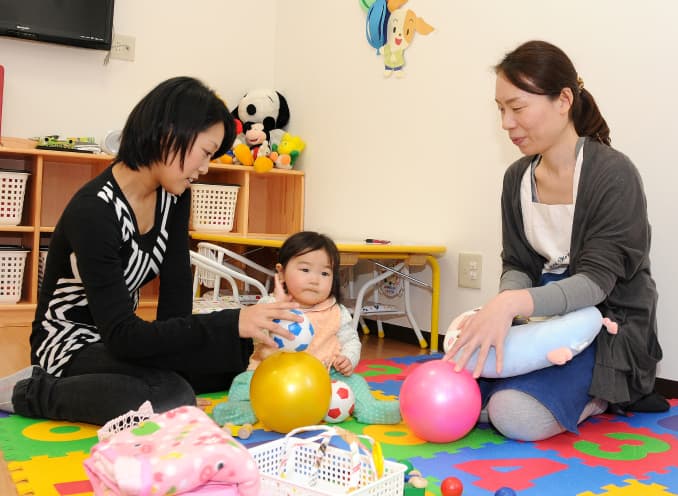 保育室で保護者と保育スタッフが幼児と一緒に遊ぶ様子。カラフルなマットの上でボール遊びをしている