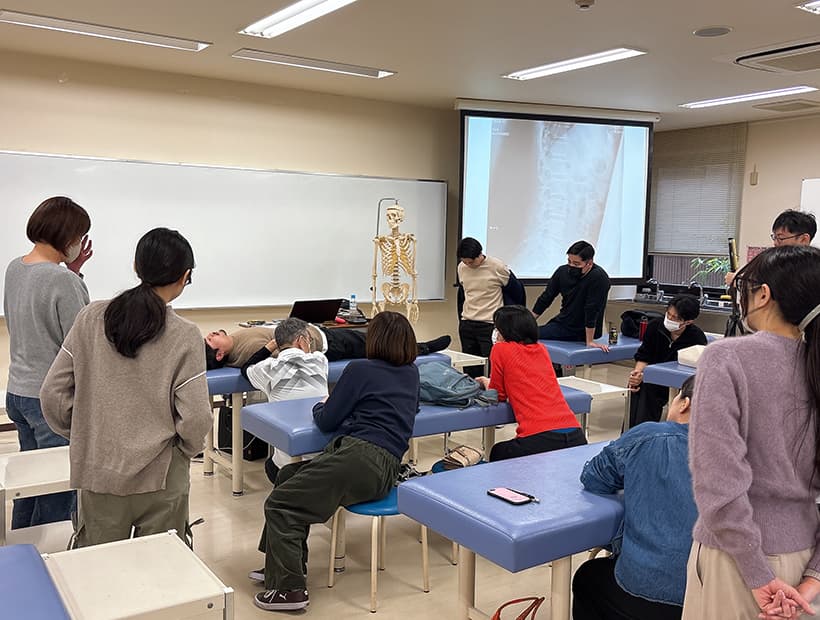 教室で横になった学生の身体を教材に、骨格模型やX線画像を用いて授業を行う様子