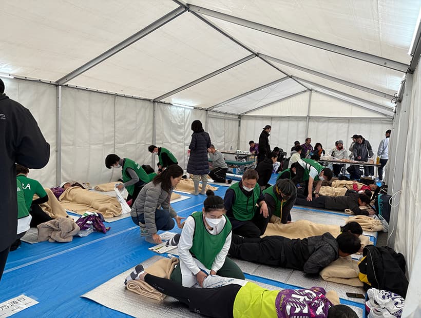 イベント会場のようなテント内で、多くの学生が参加者に対してストレッチやケアを行う実技指導の様子