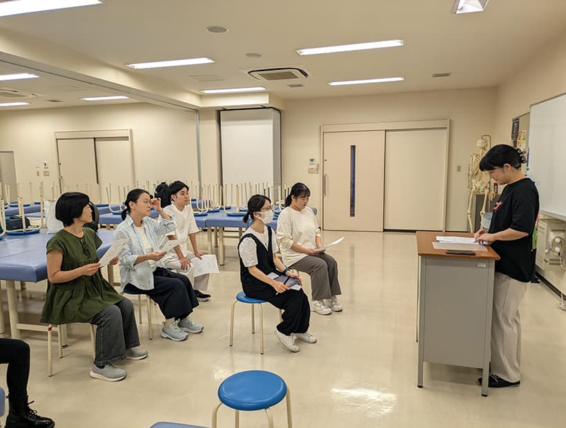 机に資料を置きながら、立って説明を行う講師と、それを座って聴講する学生たちの様子