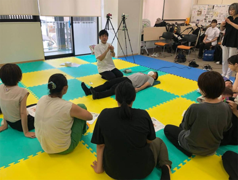 実習室で指導者の話を聞きながら、仰向けになった参加者を囲んで学ぶ学生たちの様子