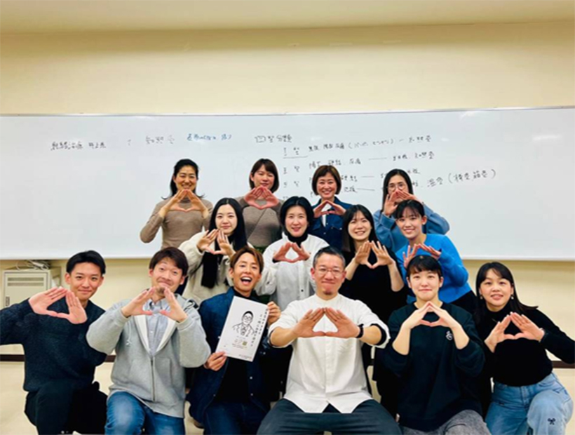 教室内で集合写真を撮る学生たち。全員が手で三角形を作ってポーズを取っている様子