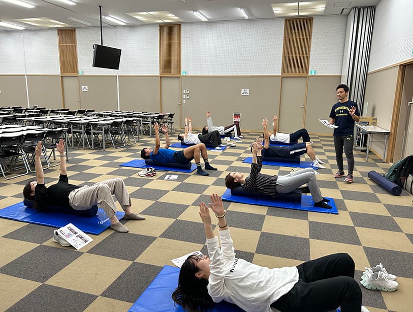 室内で指導者の指示に従い、ストレッチマットの上で運動を行う学生たちの様子